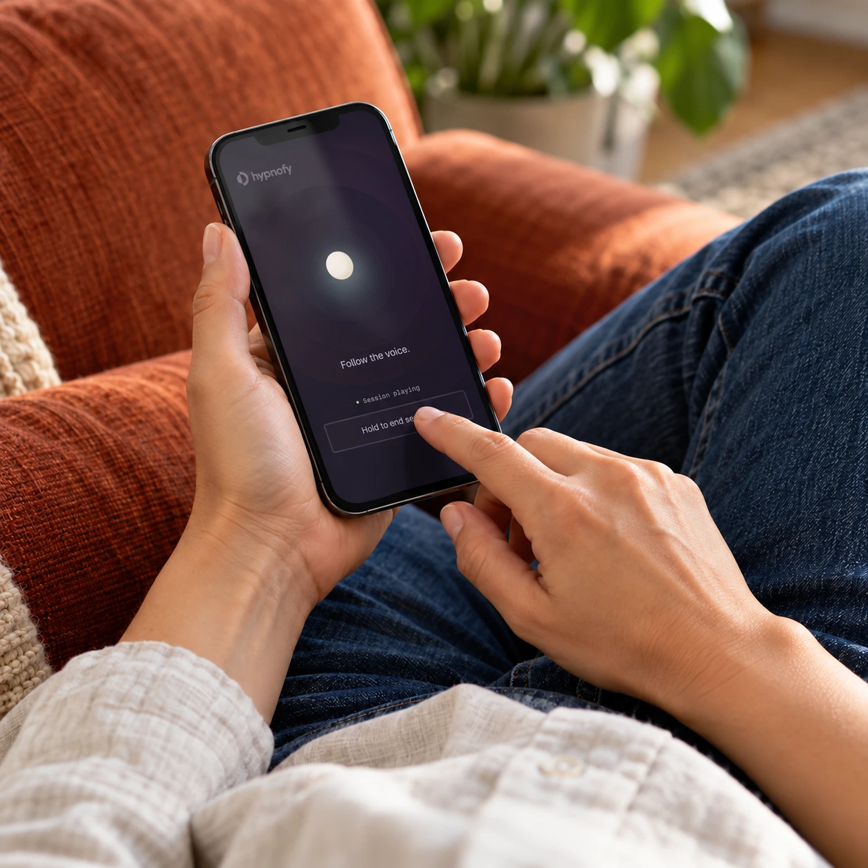 Hands holding a phone showing the Hypnofy hypnosis session while resting on a terracotta sofa.